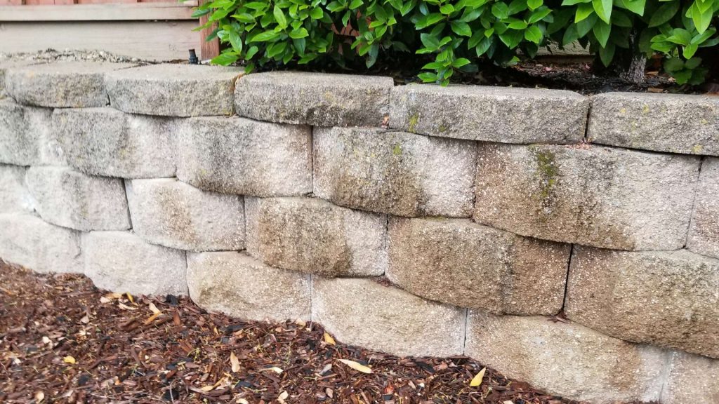 A close-up of a retaining wall made of stacked stone blocks in a garden. The wall supports a raised area with green shrubs, and the ground in front is covered in brown mulch.