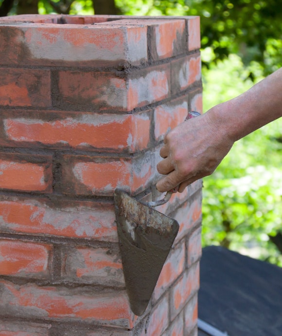 A person's hand is using a trowel to apply mortar to red brickwork, constructing a pillar. The bricks are neatly stacked, and the scene is set outdoors with greenery in the background.