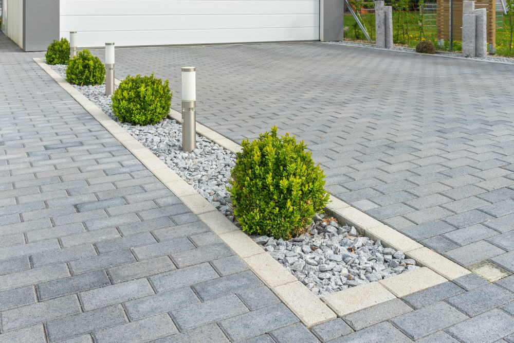 A neatly arranged driveway with grey pavers features a central strip of white gravel and green shrubs. Modern lamp posts are evenly spaced along the strip, leading to a white garage door.