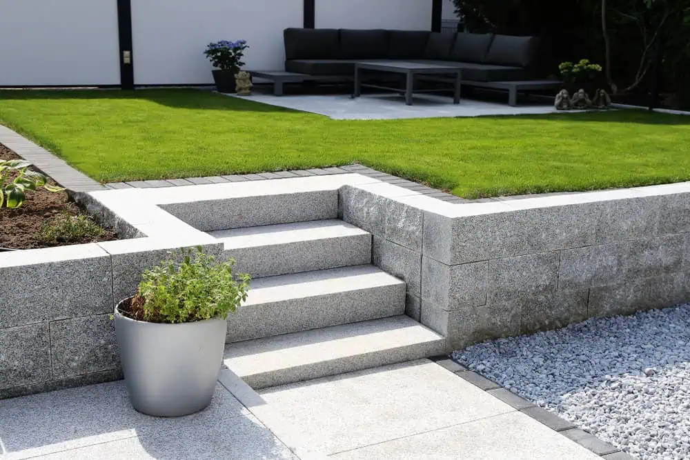 A modern backyard with a grey stone patio and steps leading to a raised grassy area. A silver planter with greenery sits on the patio. In the background, there's a dark outdoor sofa set on a concrete pad, next to plants and decorative statues.