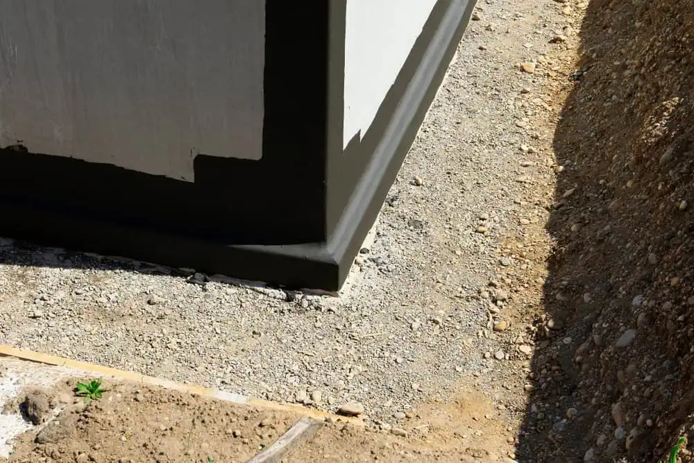 A corner of a building foundation coated with black waterproofing material. The surrounding area consists of gravel, and a trench filled with dirt and rocks runs along the right side. The image highlights foundation protection and drainage preparation in ongoing construction or maintenance work.