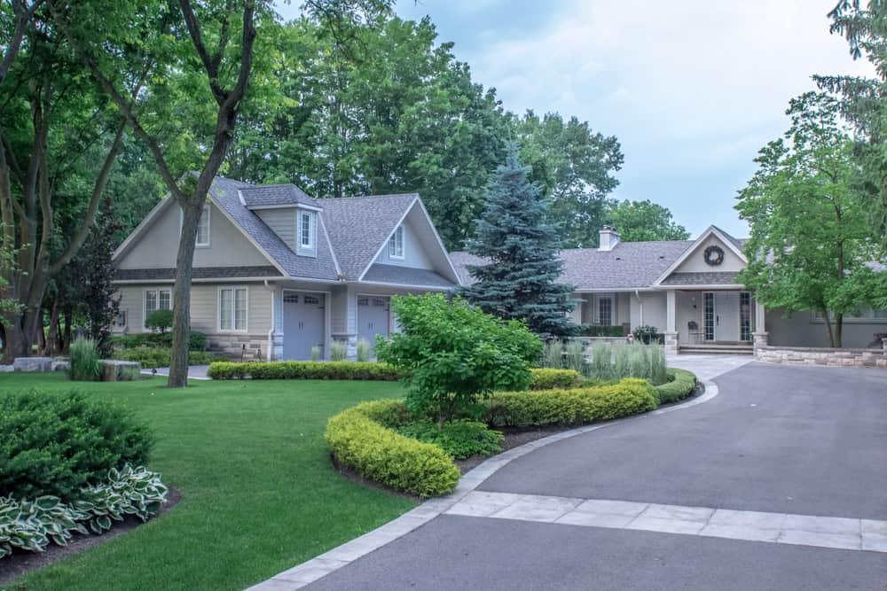 A scenic image of a suburban house surrounded by lush greenery. The foreground features a curved driveway with manicured lawns, shrubs, and trees. The house has a grayish exterior with multiple gables and large windows.