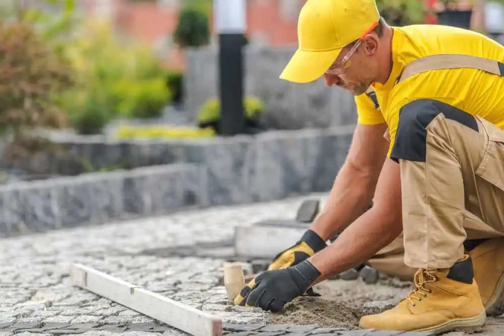 A person wearing a yellow cap and shirt, along with beige overalls and gloves, is kneeling on a cobblestone path. They are smoothing sand with a trowel. Greenery and stone steps are visible in the background.