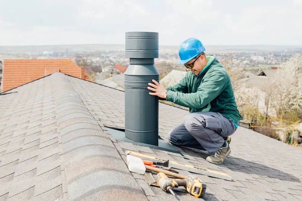 A worker in a blue helmet and green jacket kneels on a rooftop, installing a metal chimney. Various tools lie nearby on the brown shingles. In the background, a residential area and landscape are visible.