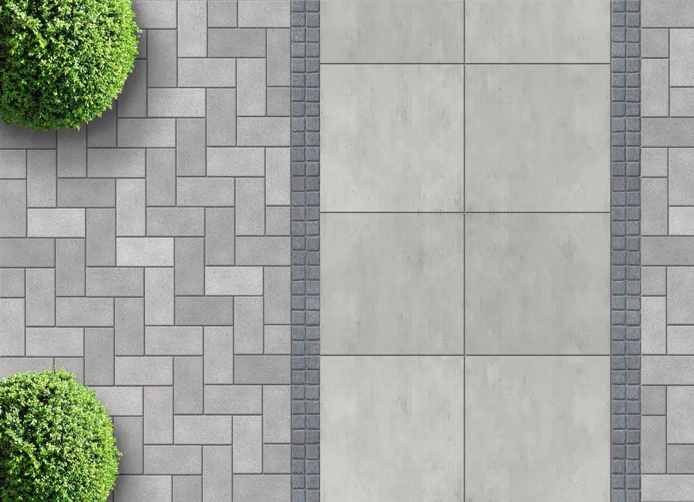 Aerial view of a walkway with large rectangular concrete slabs bordered by small grey bricks. On the left, smaller, interlocking grey tiles form a pattern with three green, round bushes placed at intervals.
