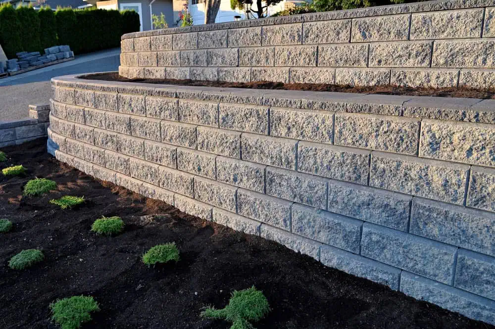 A tiered stone retaining wall with freshly planted greenery in the foreground, set against a clear sky. The stones are uniformly stacked, creating a clean, modern landscaping feature.