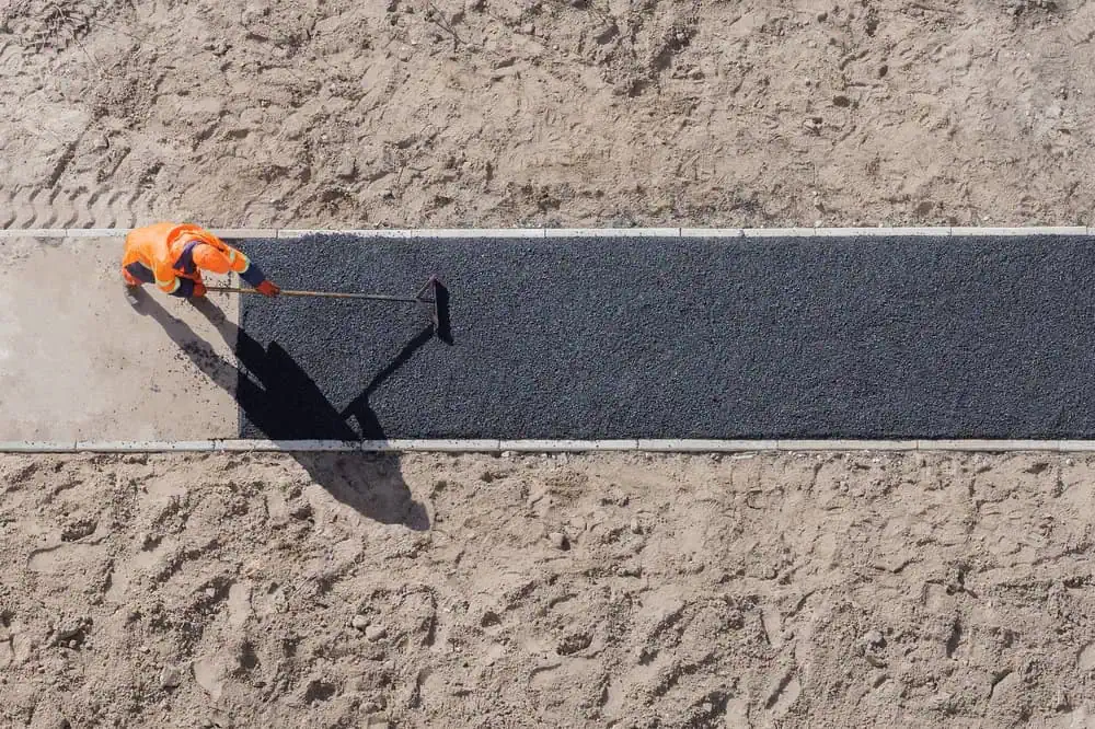 A worker in an orange safety uniform is spreading asphalt on a path in a sandy area. They are using a tool to smooth the surface, with clear, defined borders of the freshly laid asphalt. Shadows fall across the path under the sunlight.