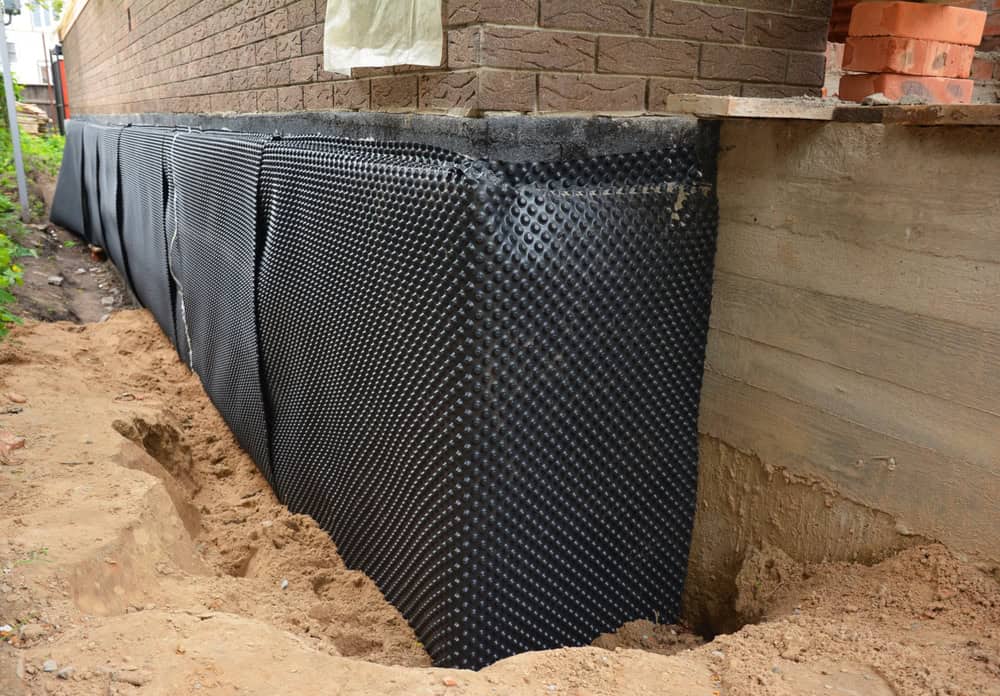 A building foundation featuring a newly installed black dimpled waterproofing membrane. The membrane is partially covered with exposed soil, indicating ongoing construction or waterproofing work. Above the foundation, the brick wall of the building is visible. The image emphasizes protective measures for foundations and proper installation of waterproofing systems.