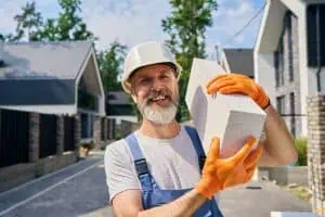 A construction worker with a beard and wearing a white hard hat, orange gloves, and blue overalls smiles while holding a large concrete block. He stands on a residential street with modern houses in the background.