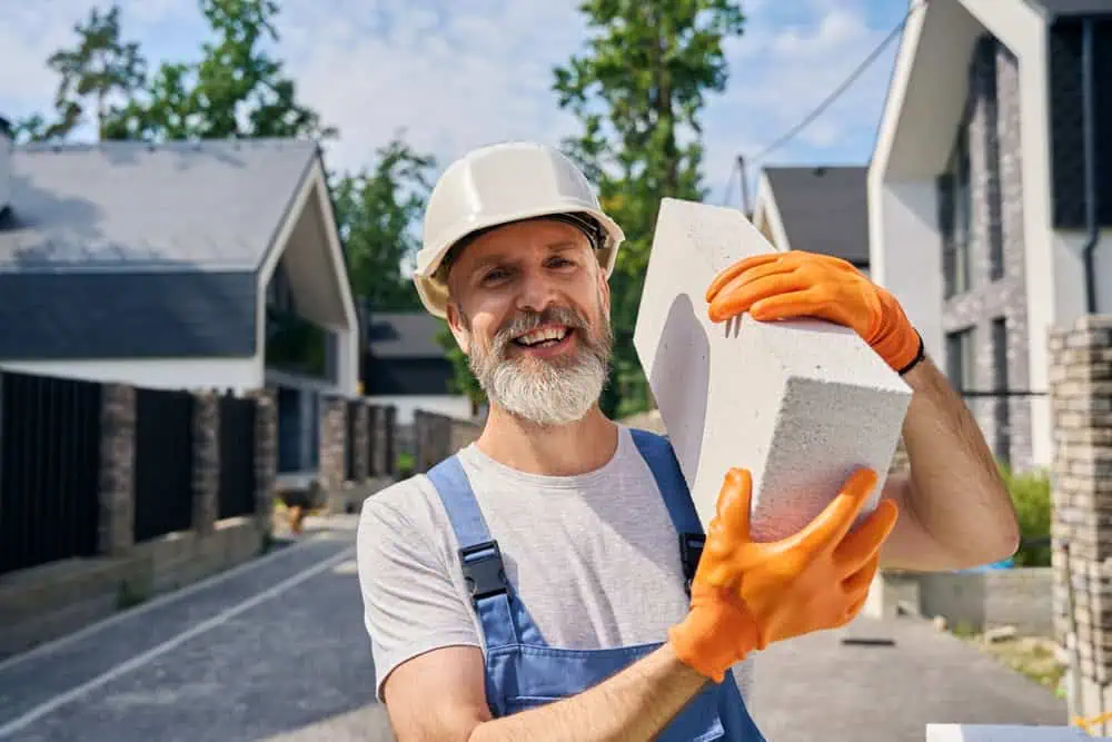 A construction worker with a beard and wearing a white hard hat, orange gloves, and blue overalls smiles while holding a large concrete block. He stands on a residential street with modern houses in the background.