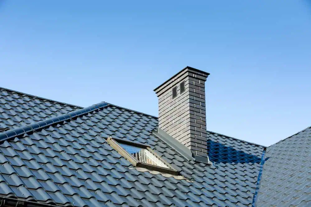 A close-up view of a house roof with blue ceramic tiles, featuring a skylight and a tall brick chimney against a clear blue sky.