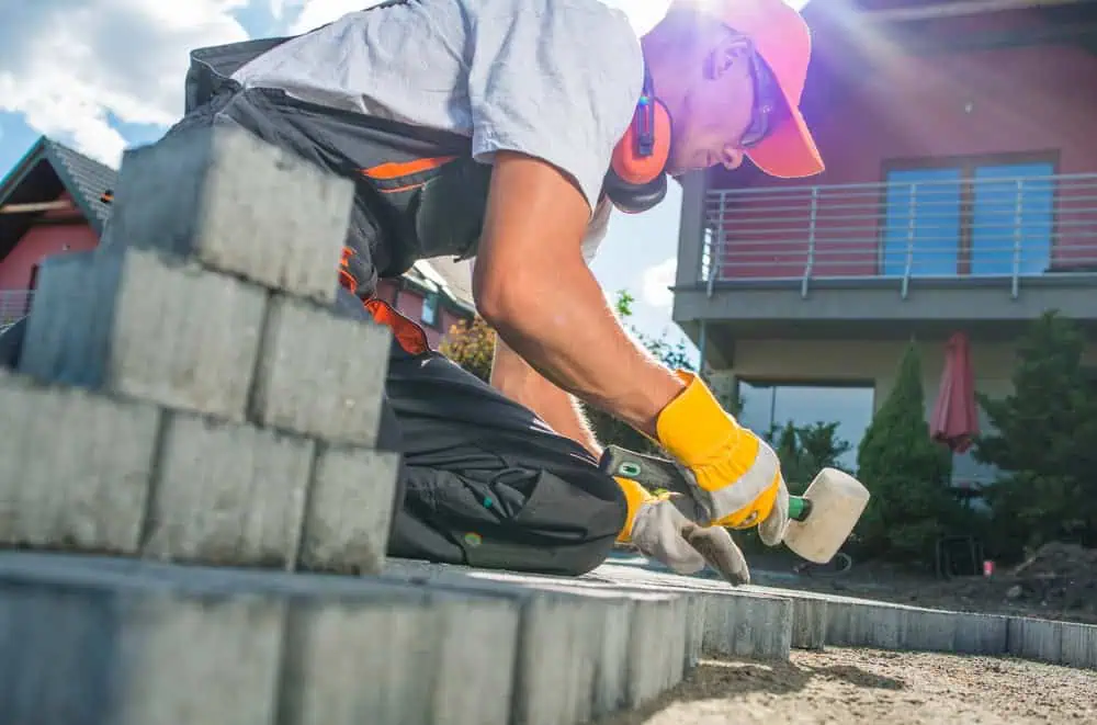 A construction worker in protective gear lays concrete blocks outdoors. Sunlight filters through the clouds, illuminating the scene. In the background, there's a modern house with a balcony and garden.