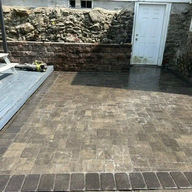 A stone-paved patio area with a white door on a stone wall. A broom and cleaning supplies are on the wooden deck to the left. The ground appears wet, suggesting recent cleaning.