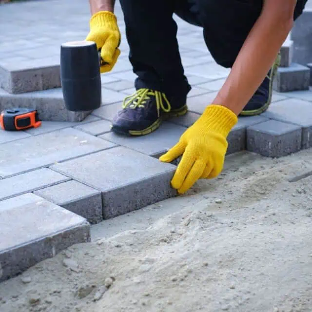 A person wearing yellow gloves and black shoes with green laces is laying gray paving stones in a sand base. They hold a rubber mallet, and a measuring tape is nearby.