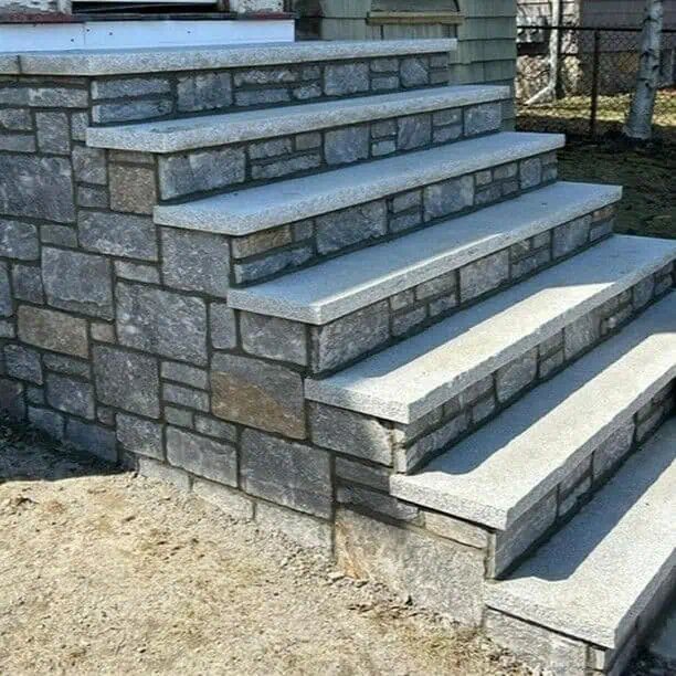 Stone steps leading up to a porch, featuring a pattern of rectangular gray stones. The steps are bordered by a stone wall on one side. The ground is bare, with a glimpse of a fenced area in the background.