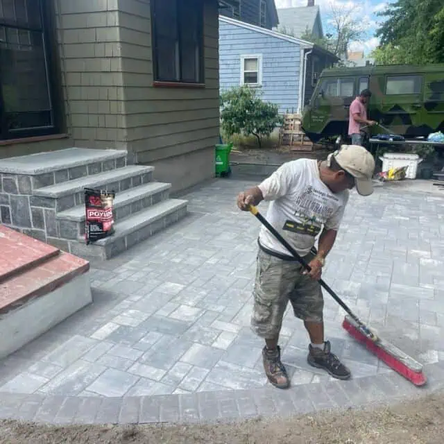 A worker sweeps a newly laid brick patio in front of a house. Bags of construction material and tools are visible. Another person stands near a green vehicle in the background. Steps lead to the house's entrance.
