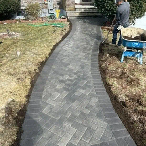 A newly paved walkway made of interlocking gray bricks curves through a garden in front of a house. Two workers are visible; one kneels on the ground while the other stands beside a wheelbarrow filled with tools and materials.