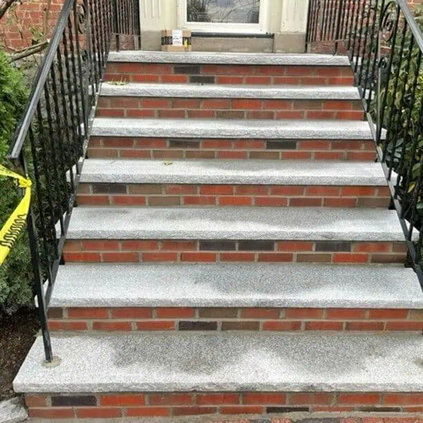 Brick and stone outdoor stairs with black metal railings on both sides. A yellow caution tape is seen on the left railing. The stairs lead up to a doorway of a red brick building. Some plants are visible on the left side.