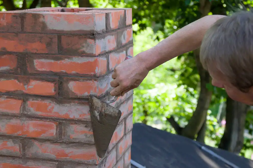 A professional mason constructing a brick chimney in Middlesex County, Massachusetts. The worker is carefully laying bricks in a precise pattern, applying mortar with a trowel, and wearing protective gloves and workwear. Scaffolding or a rooftop setting is visible, highlighting the technical skill required for safe and durable chimney construction. The image emphasizes quality craftsmanship in residential masonry projects.