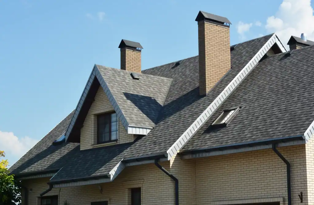 The gray shingled roof and brick chimneys of a multi-story house are shown on a sunny day.