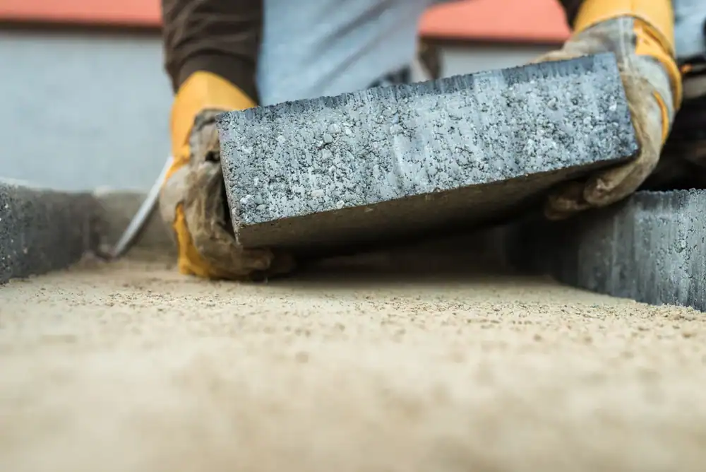 A professional construction worker laying concrete paving blocks in Middlesex County, Massachusetts. The worker is aligning interlocking concrete blocks on a prepared base of sand and gravel, using tools such as a rubber mallet and level to ensure even placement. The surrounding area shows a partially completed paved surface, highlighting careful craftsmanship in creating a durable and visually appealing walkway or patio.