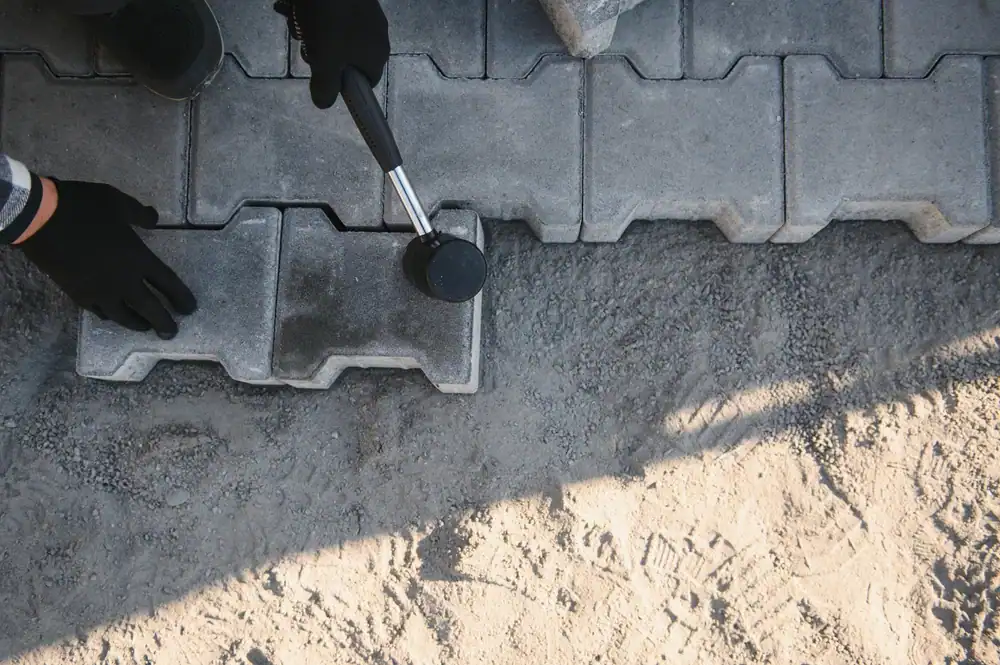A team from Academy Masonry and Construction installing paving stones in a residential outdoor area. Workers are arranging interlocking stones on a leveled base of sand and gravel, ensuring proper alignment and spacing. Construction tools, such as rubber mallets and measuring tapes, are visible, and the surrounding backyard or patio area is partially landscaped. The image emphasizes professional craftsmanship in creating a durable and visually appealing paved surface.