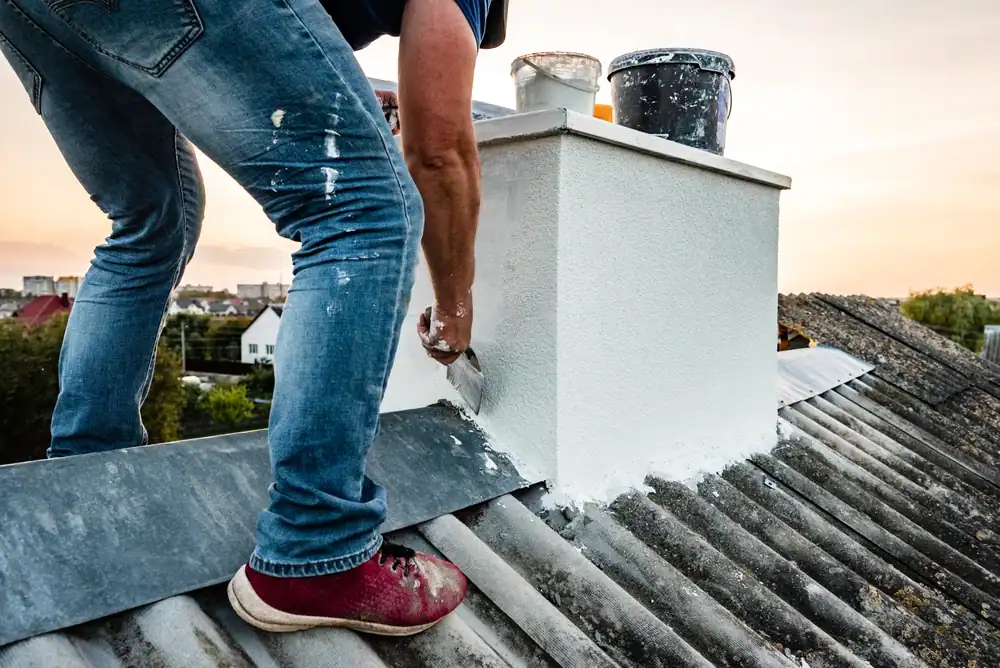 A professional masonry contractor performing chimney waterproofing and sealing on a residential rooftop. The worker is wearing safety gear, including gloves and a helmet, while applying a waterproof sealant to the brick chimney. The roof is made of asphalt shingles, and the surrounding residential neighborhood is visible in the background. The image highlights the careful application of protective materials to prevent water damage and enhance the durability of the chimney.