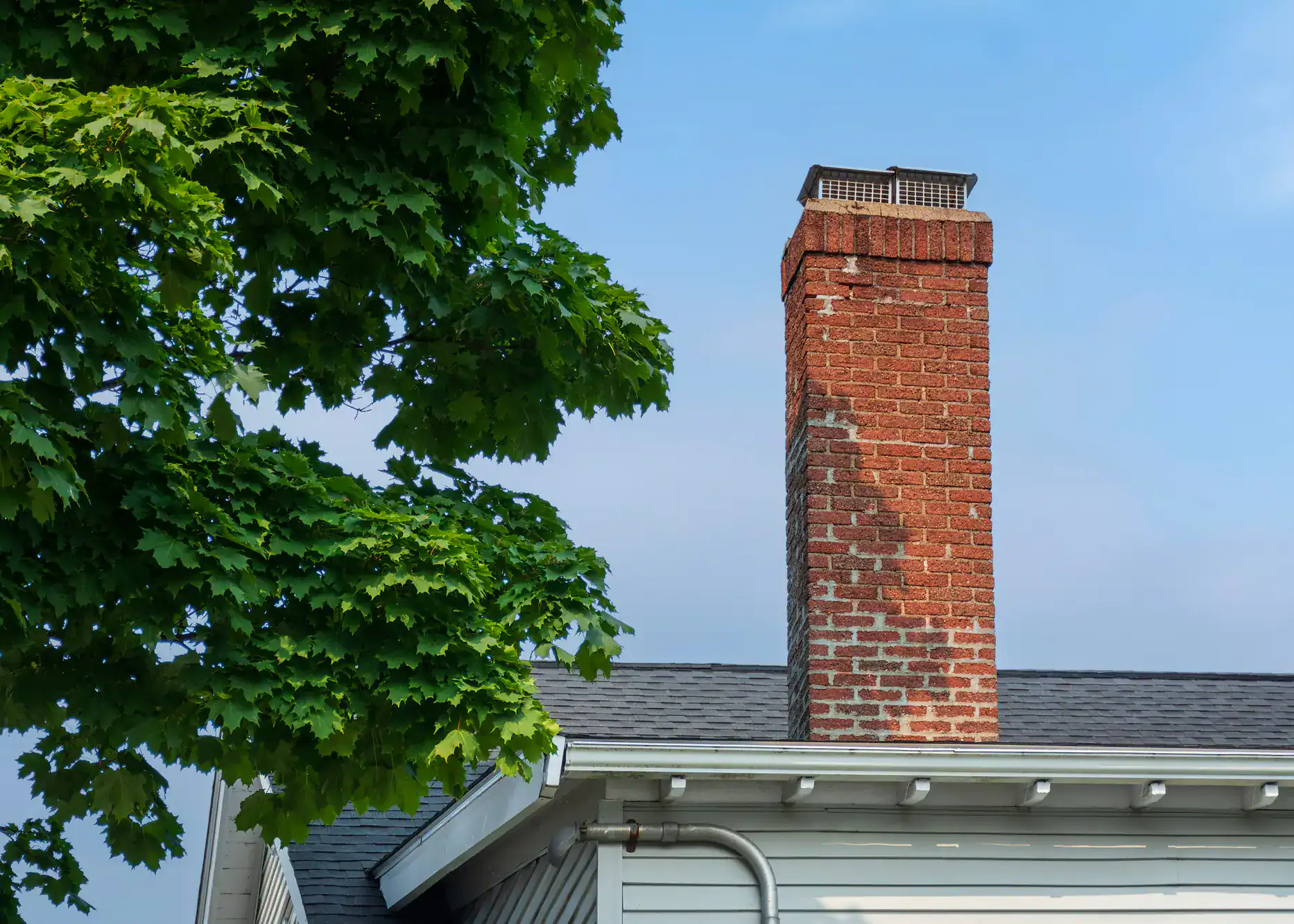 A sturdy brick chimney on a rooftop with a mature tree visible behind it, showcasing quality masonry work and outdoor structural integration by Academy Masonry