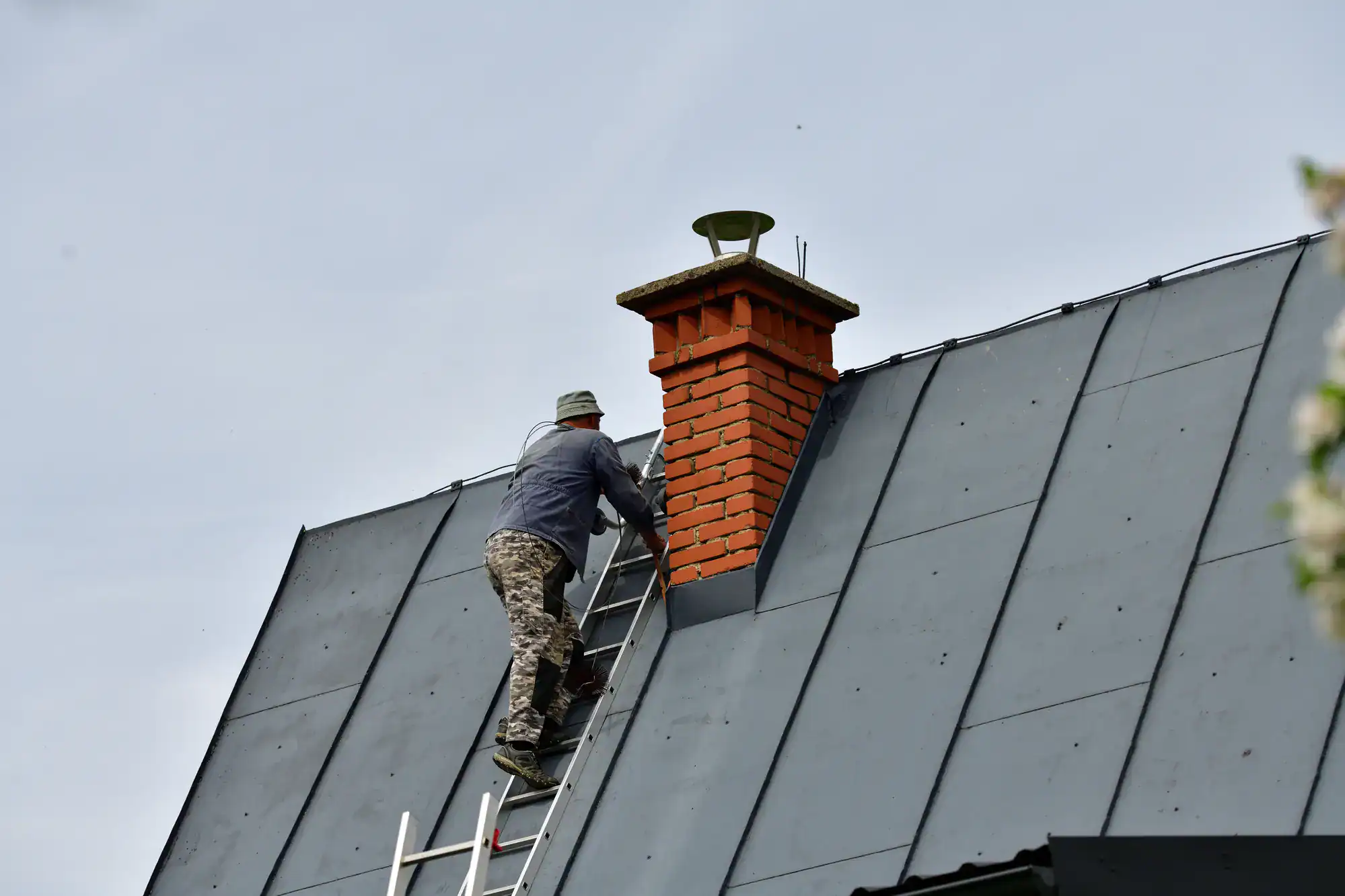 Professional mason from Academy Masonry repairing a brick chimney on a residential roof, wearing safety gear and using specialized tools to restore and maintain the chimney structure.