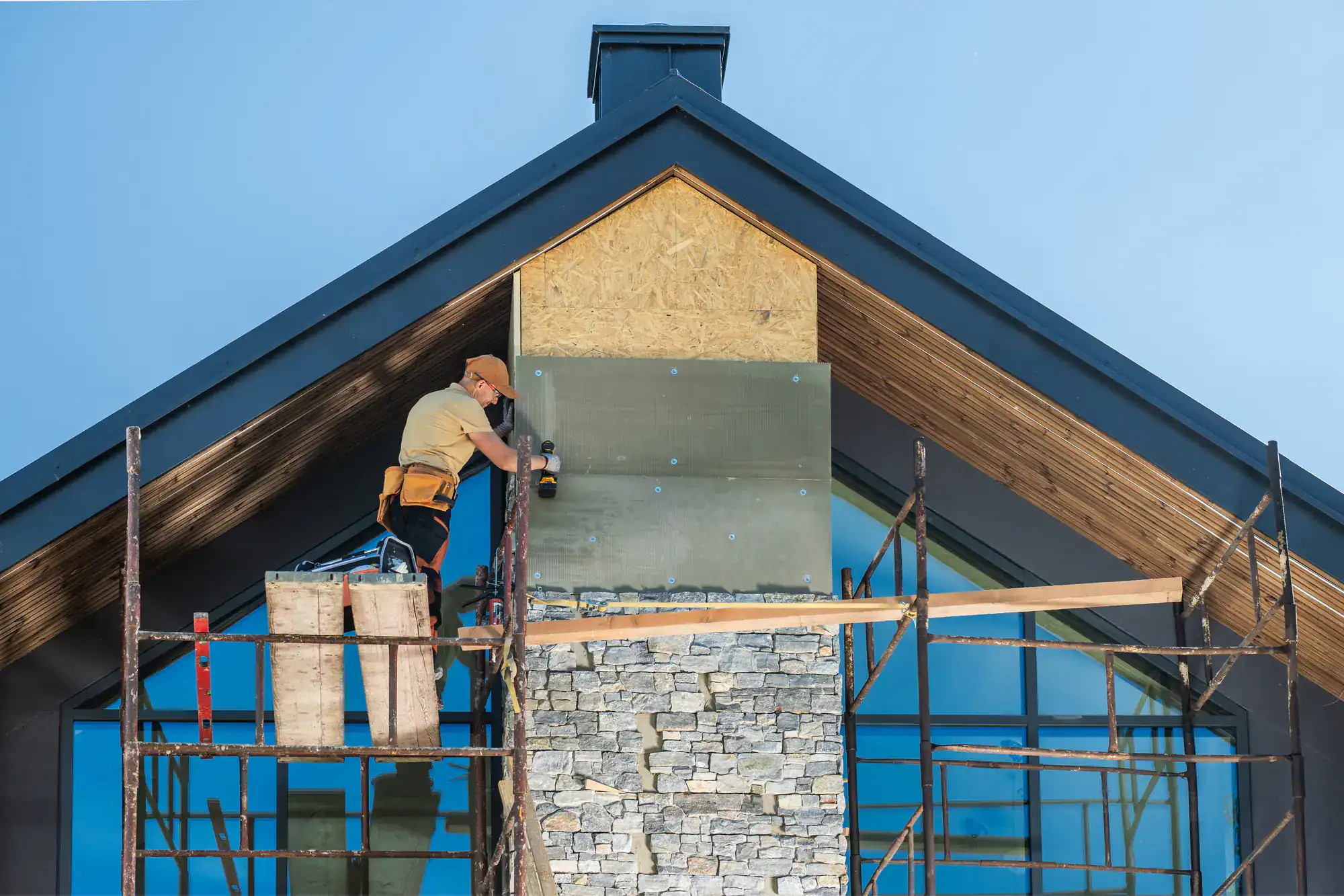 Construction worker wearing a hard hat and safety harness, standing on a scaffold while working on a roof. The worker is handling masonry materials as part of a roofing project for Academy Masonry, with tools and bricks visible on the scaffold and a partially completed roof in the background.